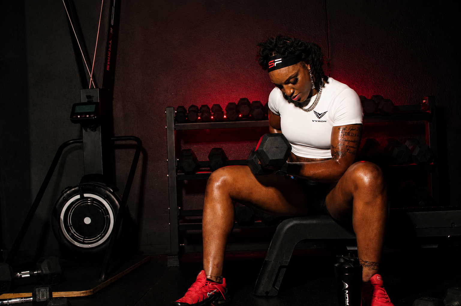 A woman in a Vyron white athletic top and red sneakers sits on a bench in a dark gym, holding a hex dumbbell. A rowing machine is visible on the left, and red lighting illuminates the background.
