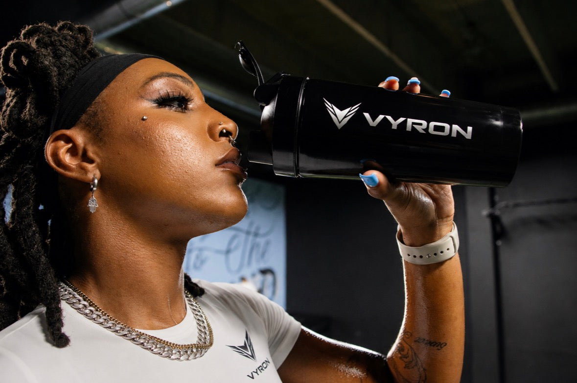 A woman in a gym setting drinking from a black Vyron shaker bottle. She is wearing a white Vyron t-shirt, a silver chain necklace, and a black headband, with blue nail polish on her hand.