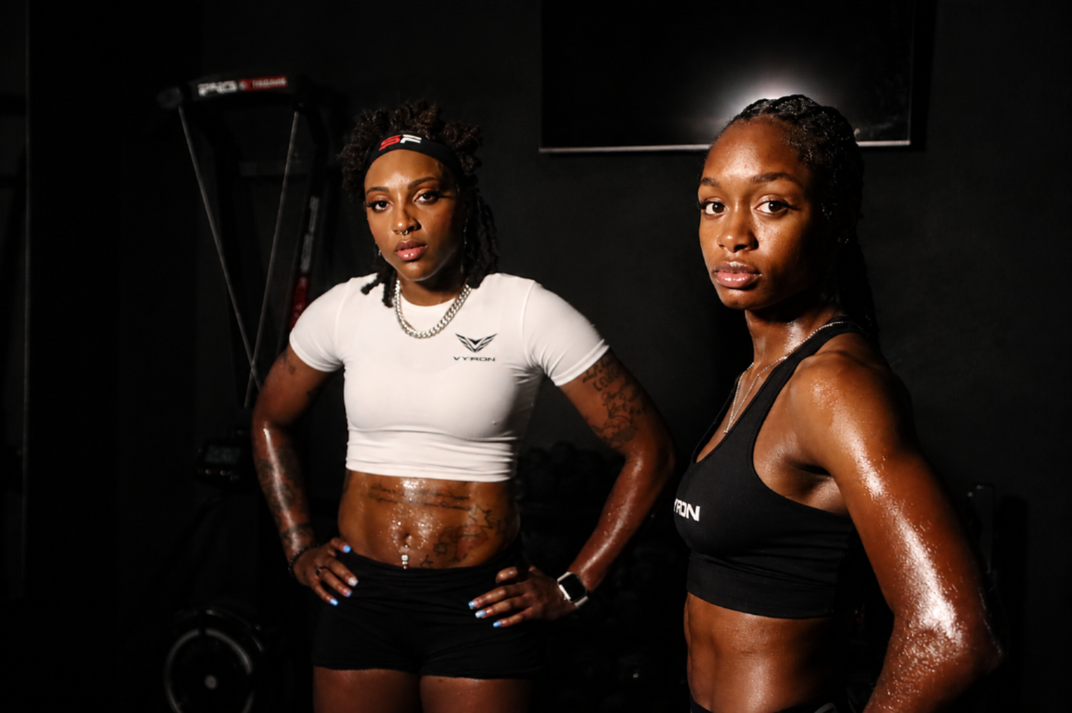 Two athletic women posing in a dark gym setting, wearing branded sports apparel: a white cropped T-shirt with a Vyron logo on the left and a black sports bra with a visible white Vyron logo on the right.